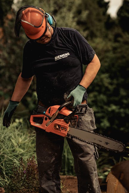 A gardener wearing a wide-brimmed hat and dark clothing is trimming dense green hedges with long-handled pruning shears, focusing on shaping the rounded foliage. The garden features a lush hedge compo